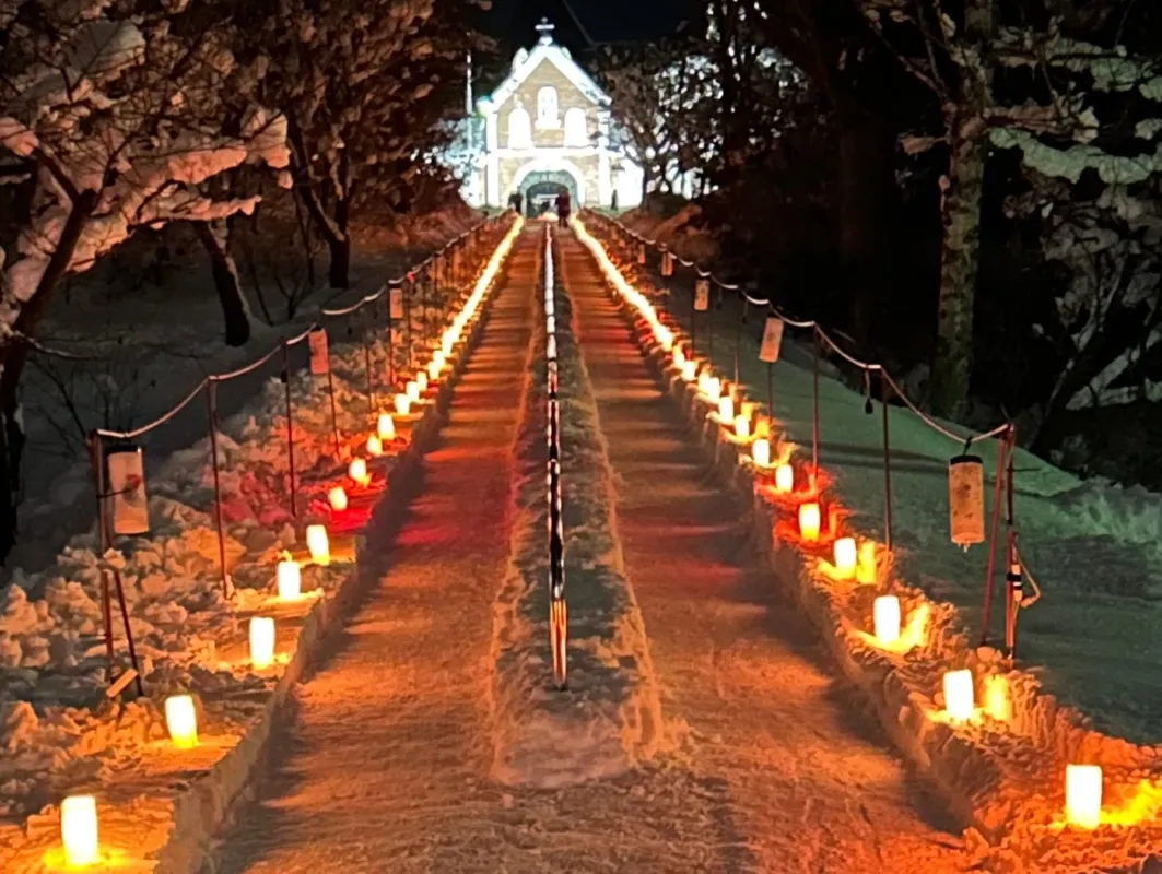 雪景色の中、灯りが並んだ小道を進むと教会の建物が見える写真です。