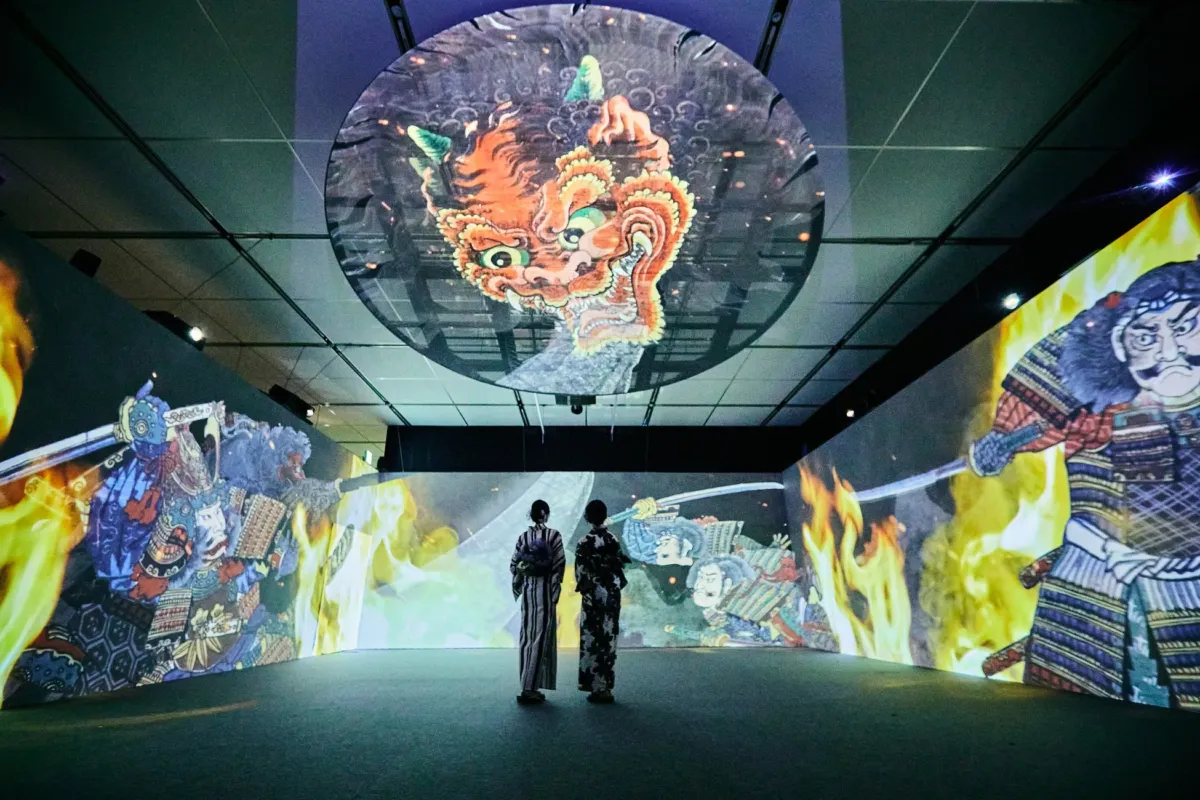 二人の女性が、壁一面に武士の絵が描かれた部屋に立っている様子。天井には大きな鬼の顔のプロジェクションマッピングが映し出されている。