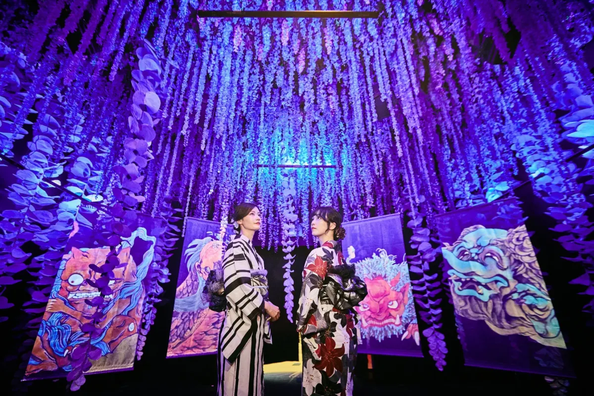 2人の女性が着物を着て、紫色の装飾が施された空間に立っています。背景には、日本の伝統的な絵画が飾られています。