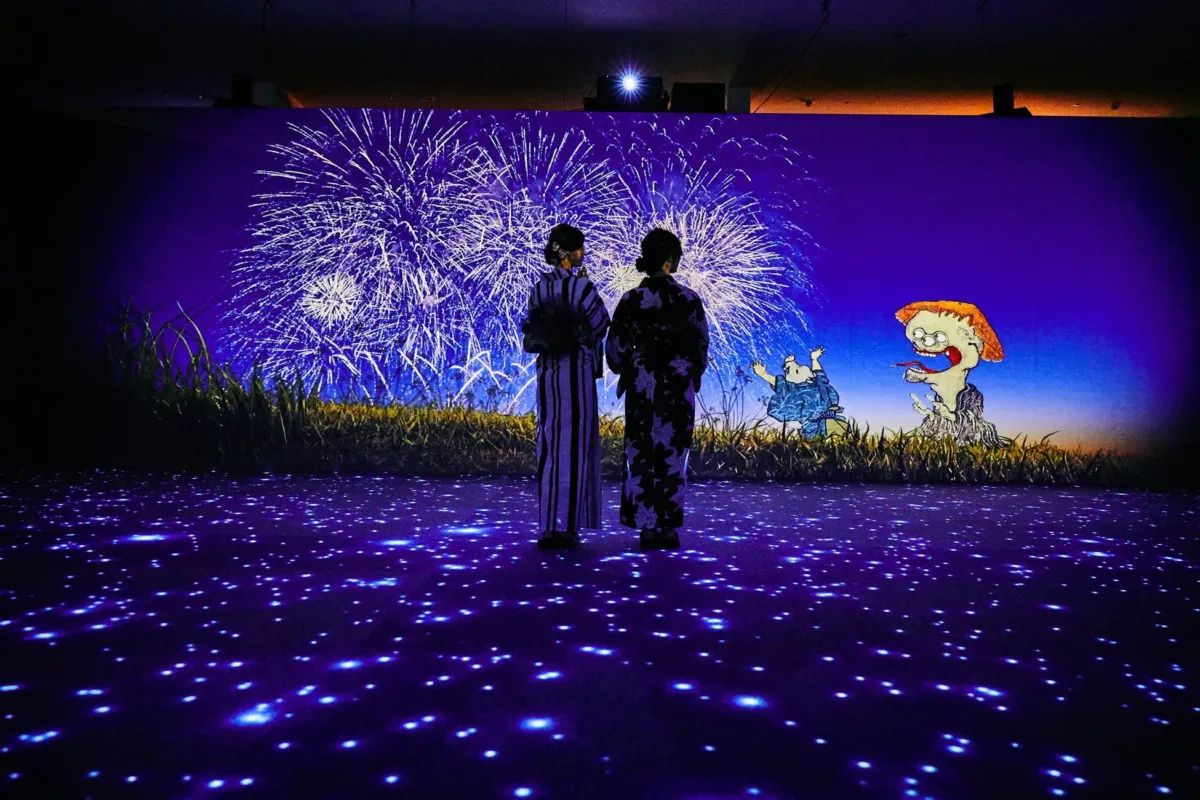 花火のプロジェクションマッピングを見ている二人の女性。足元には光の演出が施され、背景には妖怪のようなキャラクターも描かれている。