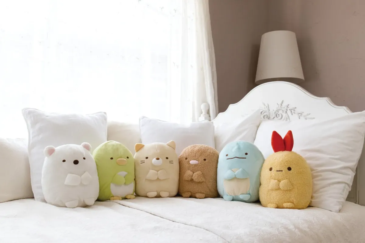 白いベッドの上に、すみっコぐらしのぬいぐるみたちが並んで置かれている様子を写した画像。それぞれのキャラクターが可愛らしく、様々な色と形で表現されている。背景には白いカーテンと枕があり、柔らかい雰囲気を作り出している。
