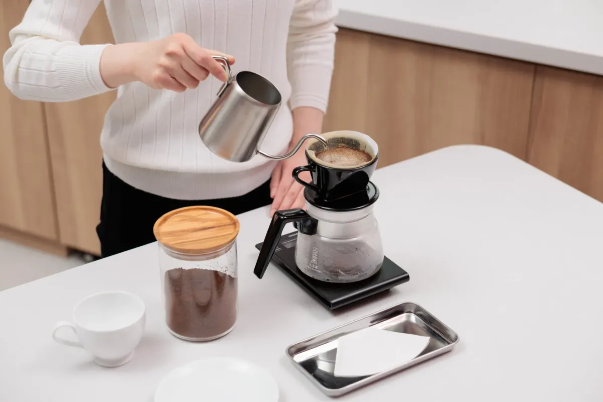 コーヒーを淹れている様子。女性がケトルからドリッパーに湯を注いでいる。コーヒー豆、カップ、サーバー、ペーパーフィルターなど、コーヒーを淹れるための道具が写っている。