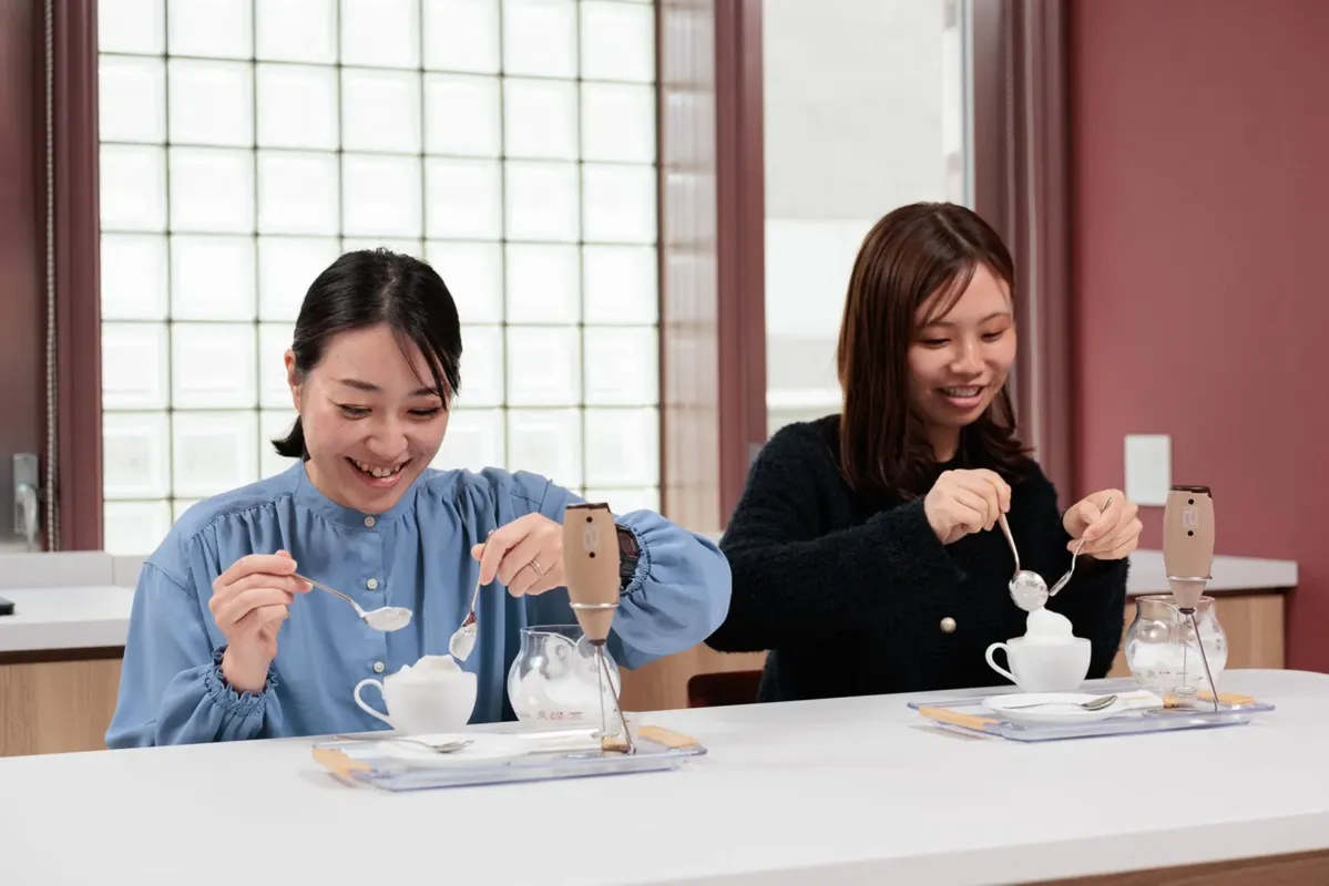 2人の女性が、テーブルで泡立て器を使って飲み物にクリームを乗せている様子。